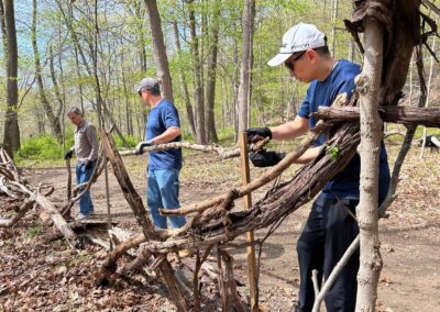 Three Men Wearing Gloves and Caps Work with Long Fallen Branches in a Forest Clearing Stacking Wood Pieces As a Barrier or Fence